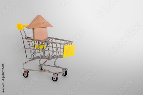 House model made of wooden cubes in a grocery cart with a yellow handle on a gray background. Side view. Copyspace