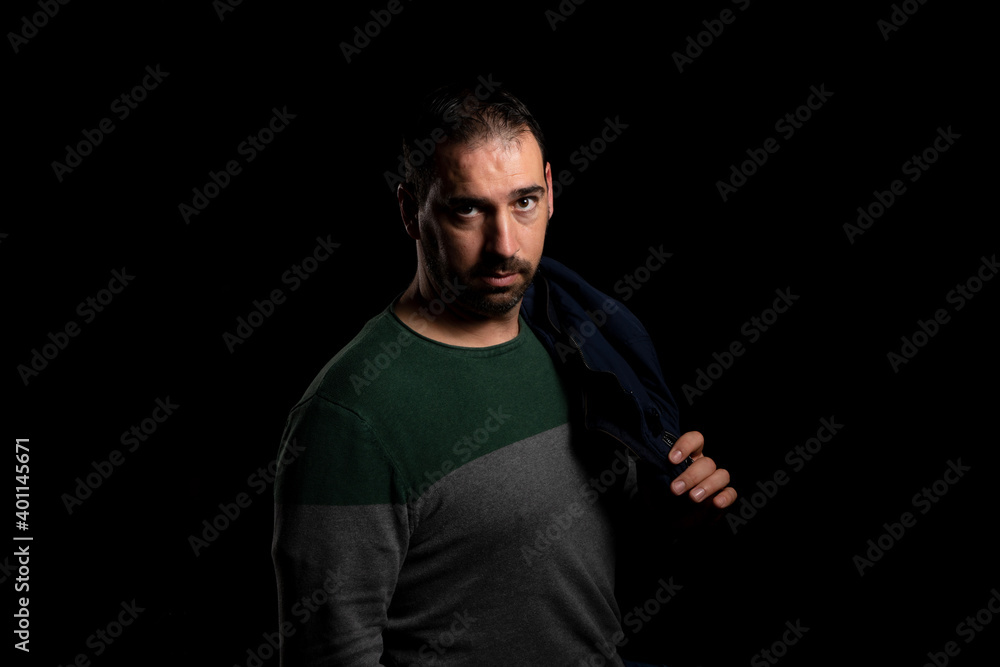 Attractive bearded man in a blue jacket slung over his shoulder posing isolated on black background