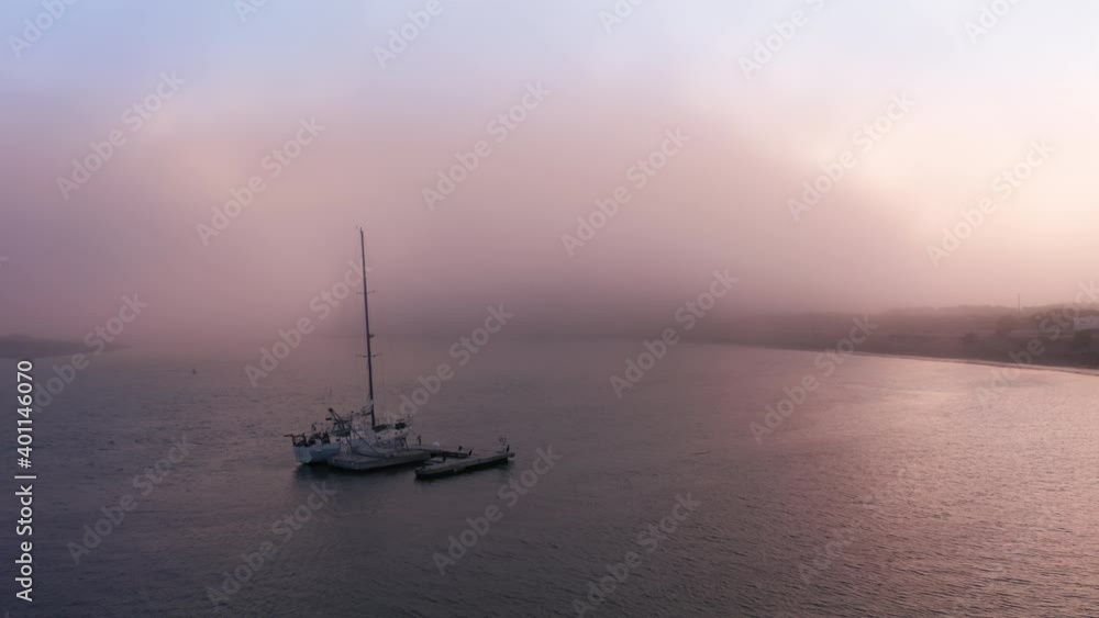 Cinematic aerial of Sailing yacht moored off Pacific ocean coast in bay before sunset or sunrise with pink and purple light coming through the fog or cloud floating above water surface. Romantic 4K Cinematic aerial of Sailing yacht moored off Pacific ocean coast in bay before sunset or sunrise with pink and purple light coming through the fog or cloud floating above water surface. Romantic 4K
