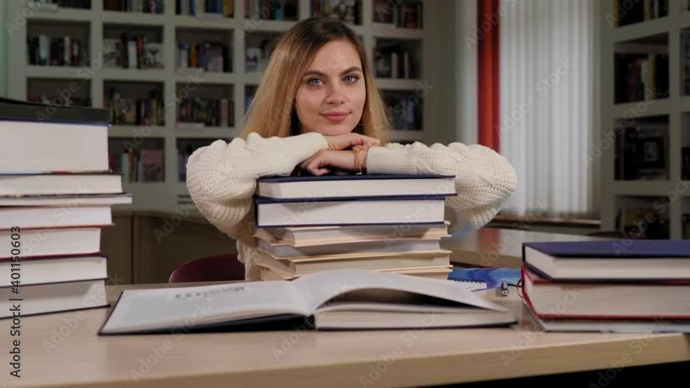 Portrait of a young beautiful girl student in a sweater and with long blonde hair in the library, she lies on a large stack of books leaning on her hands. Portrait of a young beautiful girl student in a sweater and with long blonde hair in the library, she lies on a large stack of books leaning on her hands.