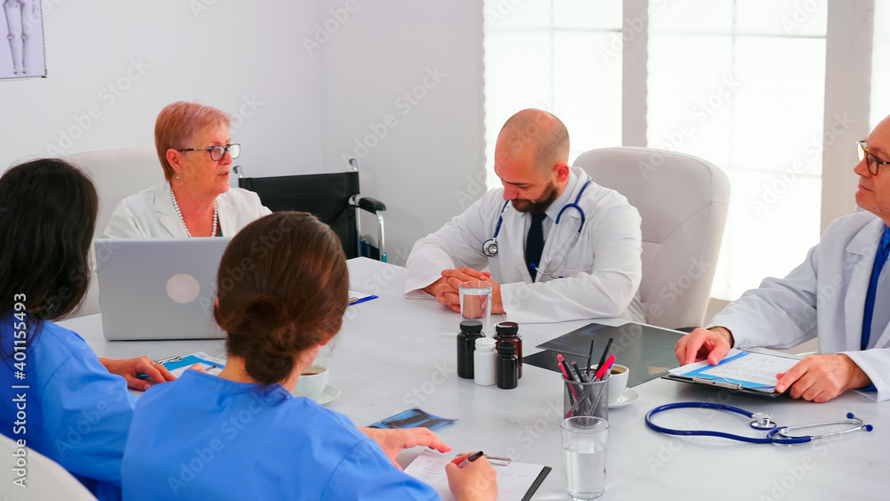 Obraz premium Mature doctor presenting health report during meeting with coworkes in hospital conference room. Clinic expert therapist talking with colleagues about disease, medicine professional