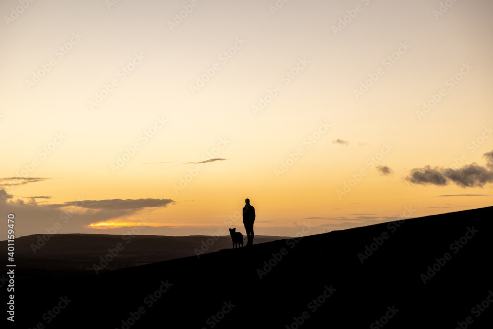 Silhouette of man and his dog looking at the sunset and enjoying the view over the countryside hill