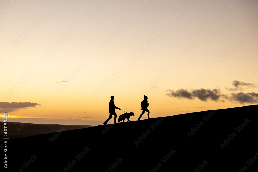 Silhouette of Man and Woman couple walking their dog at sunset on a hill with beautiful clouds and orange hue