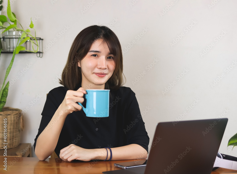 Asian woman wearing black shirt, sitting at wooden table with computer laptop and plant pot, holding blue coffee cup, smiling and looking at camera. white wall with house plant background. Asian woman wearing black shirt, sitting at wooden table with computer laptop and plant pot, holding blue coffee cup, smiling and looking at camera. white wall with house plant background.