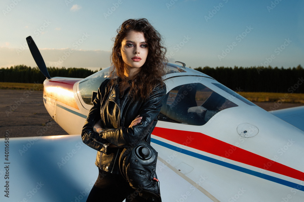 Beautiful sexy girl in a leather flight suit on a sunny day with a sunset  sky on the background of a small two-seater plane Stock Photo | Adobe Stock