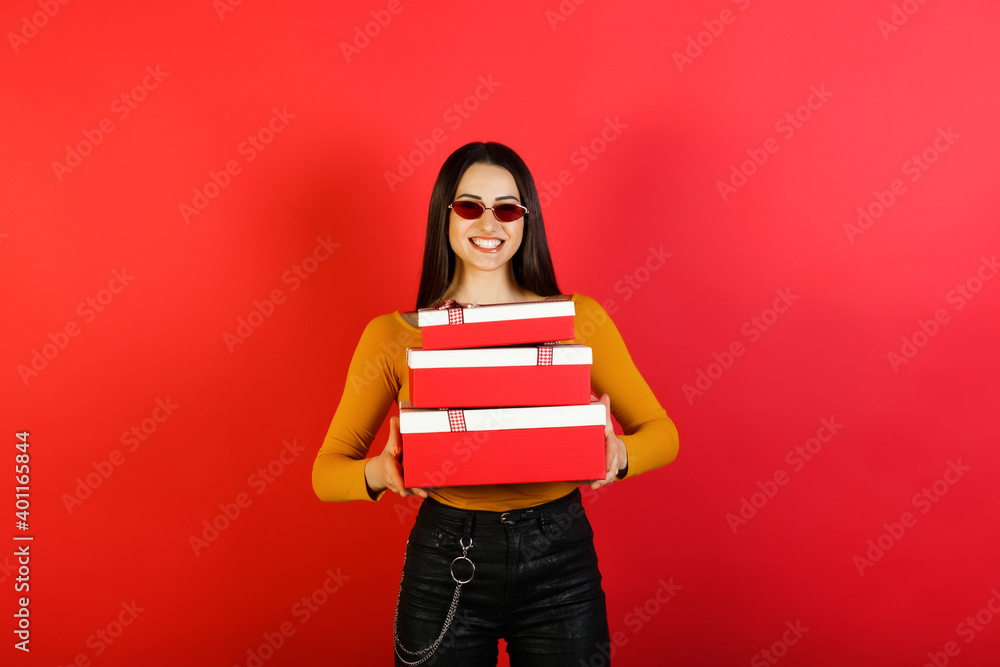 Happy stylish woman with red sunglasses holding red gift boxes and looking at the camera while standing over red background. Happy stylish woman with red sunglasses holding red gift boxes and looking at the camera while standing over red background.