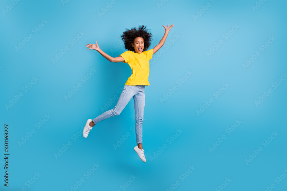 Full length photo of little cute afro american girl jump air raise hands wear jeans yellow t-shirt isolated on blue color background
