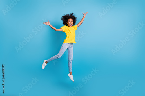 Full length photo of little cute afro american girl jump air raise hands wear jeans yellow t-shirt isolated on blue color background