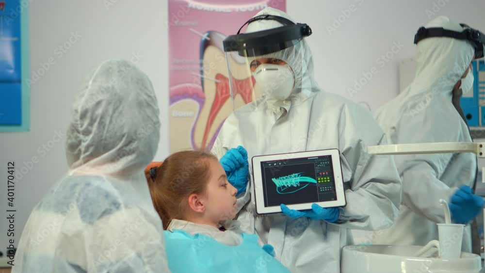 Dentist with ppe suit pointing on digital screen explaining x-ray to mother of girl patient. Medical team and patients wearing face shield coverall, mask, gloves, showing radiography using notebook Dentist with ppe suit pointing on digital screen explaining x-ray to mother of girl patient. Medical team and patients wearing face shield coverall, mask, gloves, showing radiography using notebook