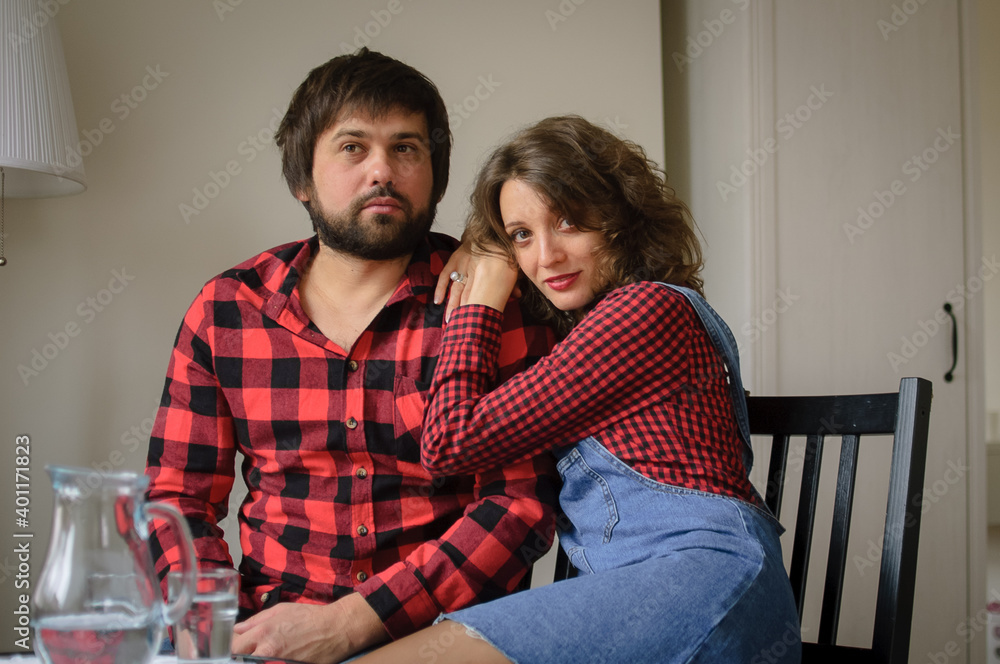 Lovely couple wearing similar red checkered shirts is hugging sitting at the table at home during celebration St. Valentine Day. Indoor portrait of boyfriend and girlfriend Lovely couple wearing similar red checkered shirts is hugging sitting at the table at home during celebration St. Valentine Day. Indoor portrait of boyfriend and girlfriend