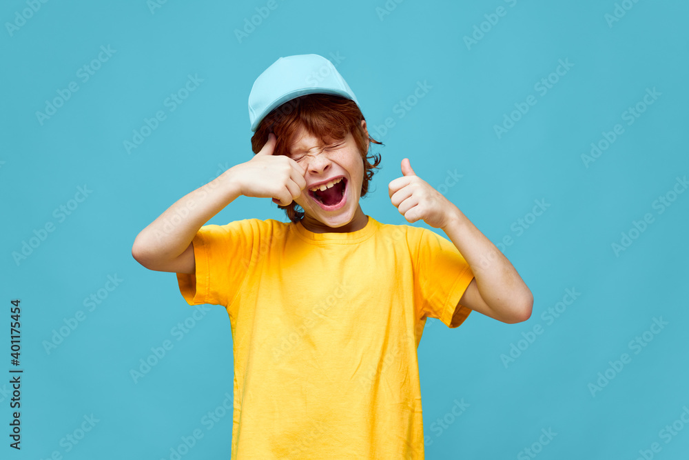 Fototapeta premium Cheerful red-haired boy with open mouth holds thumbs up yellow t-shirt cap 