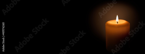  White candle on a black background. The candle flame illuminates the space around it. Banner with space for text.