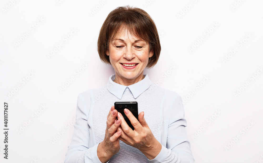 Pretty senior female in casual outfit using modern smartphone and laughing out loud while standing on white background. Old woman watching funny videos on internet. Pretty senior female in casual outfit using modern smartphone and laughing out loud while standing on white background. Old woman watching funny videos on internet.