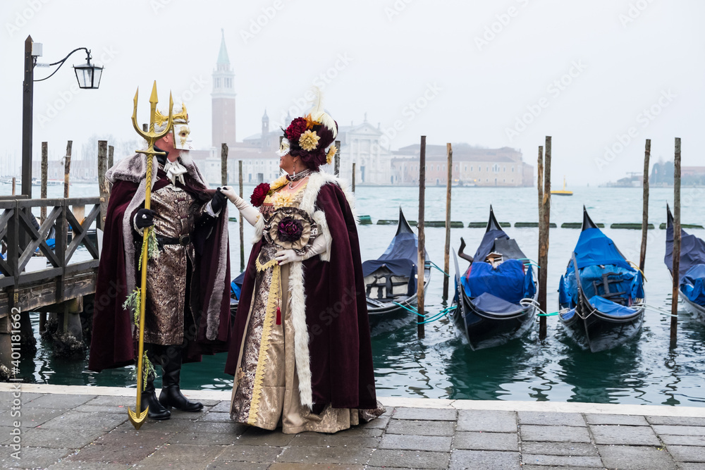 Venice, Italy - February 17, 2020: An unidentified couple in a carnival costume in front of a group of gondolas and St Giorgio's Island,  attends at the Carnival of Venice.