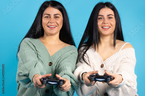 Two twin sisters with joysticks on a blue background posing, smiling and looking at the camera.