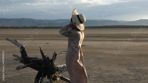 Woman in designer long loose beige dress puts on trendy straw hat standing near tree snag against mountains side view slow motion photo