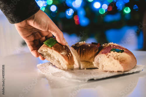 a hand picking up a piece of roscón de reyes, ring pastry roll, with an illuminated Christmas tree. Christmas concept and typical food of Spain