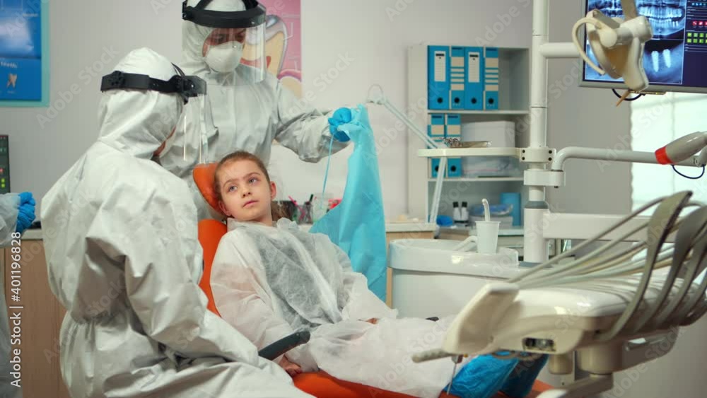 Man assistant in protective suit putting dental bib to kid before stomatological examination during covid-19 pandemic. Concept of new normal dentist visit in coronavirus outbreak wearing coverall Man assistant in protective suit putting dental bib to kid before stomatological examination during covid-19 pandemic. Concept of new normal dentist visit in coronavirus outbreak wearing coverall