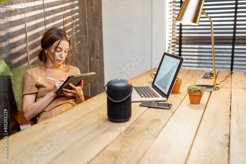 Woman working on the digital tablet at cozy home office. Smart speaker on the table. Concept of a smart home and work from home