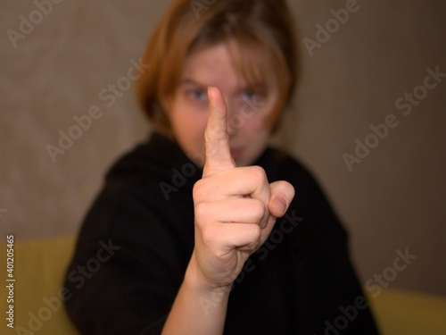 A blonde woman with black hoodie in cream background pointing her index finger at the camera 