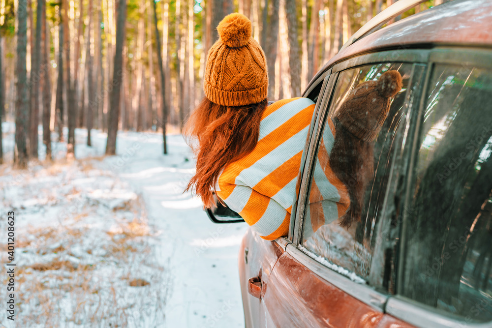 A woman in a sweater and hat looks out of the car window against the background of a snowy landscape in a winter pine forest, road trip