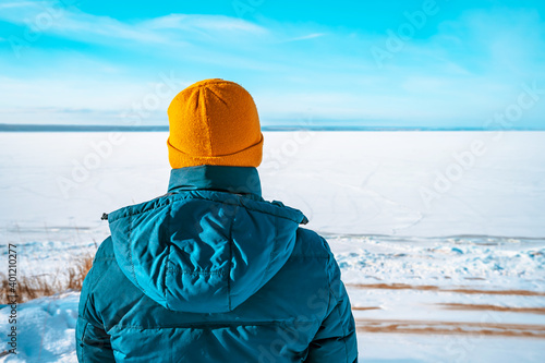 Rear view of man in a winter jacket and yellow hat stands against the background of a frozen river with a winter landscape