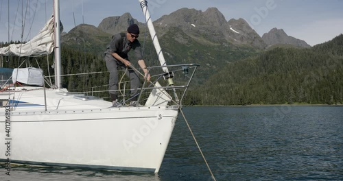 Sailing in Alaska. Man Pulling Rope at Bow of Sailboat With Scenic Coastline in Basckground, Slow Motion photo