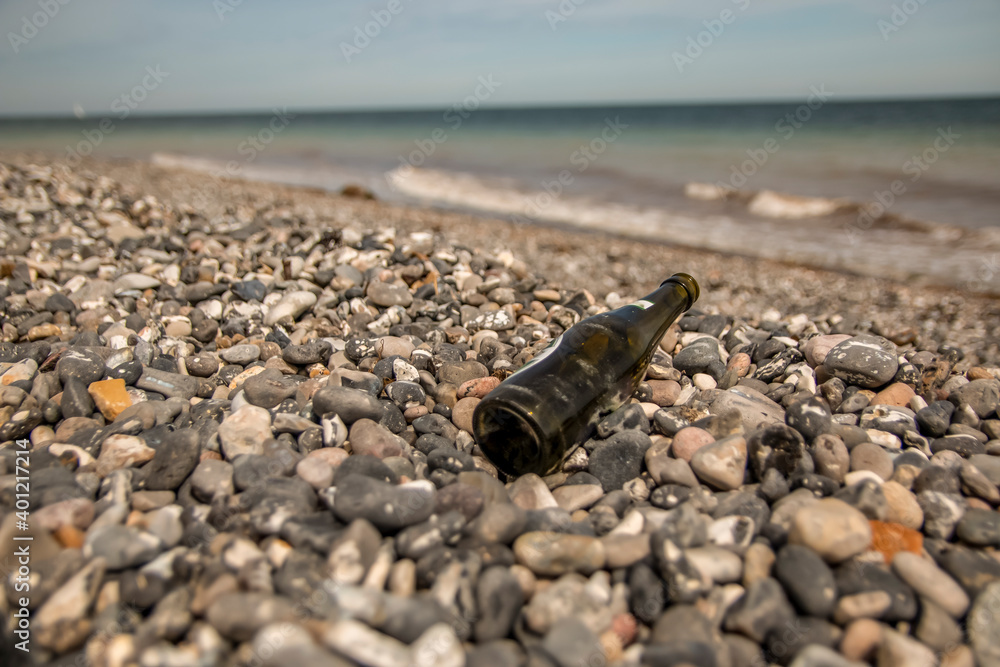A green beer bottle lies in the rocks on a beach, the sea and waves are seen in the background A green beer bottle lies in the rocks on a beach, the sea and waves are seen in the background