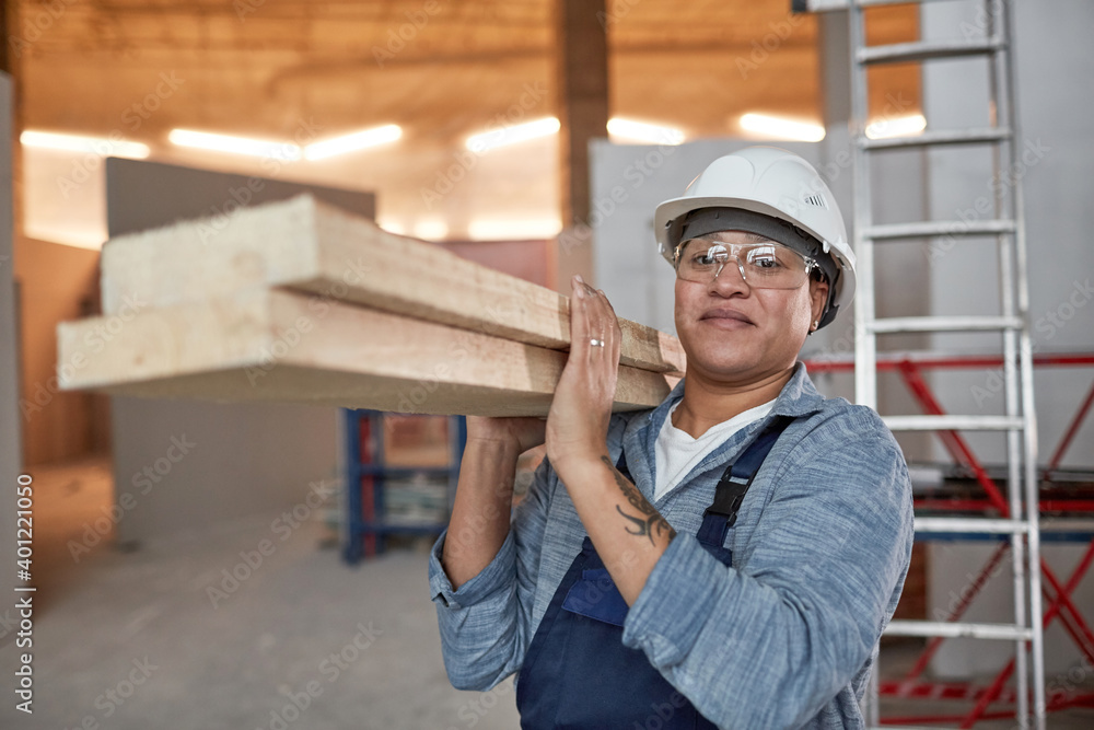 Waist up portrait of smiling female worker carrying wood boards while working on construction site, copy space Waist up portrait of smiling female worker carrying wood boards while working on construction site, copy space