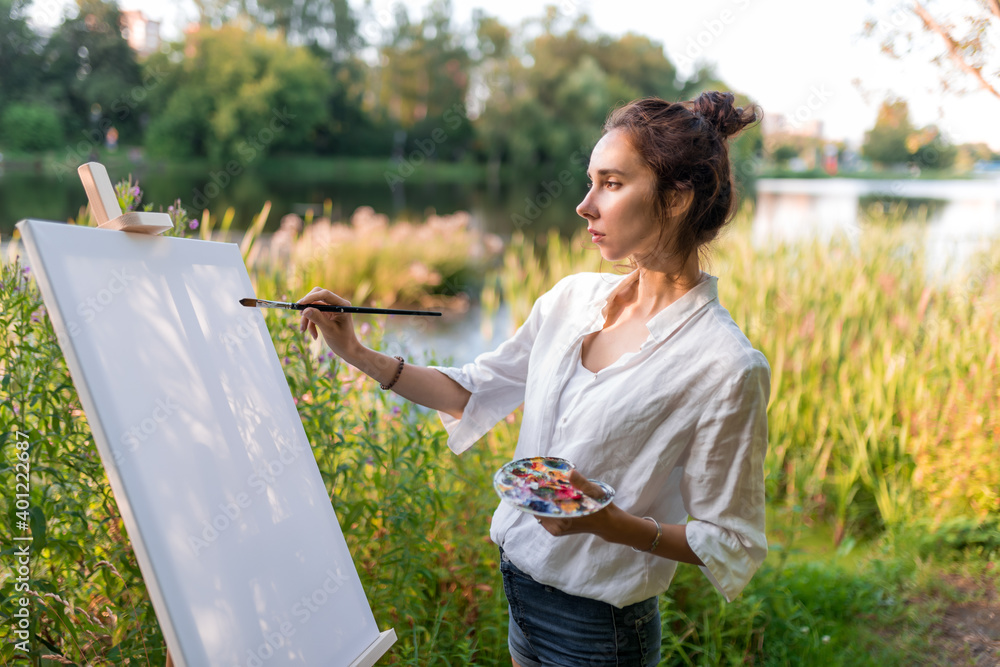 Naklejka premium beautiful girl, woman artist, in summer on shore of lake, river pond, draws picture, beginning drawing, first brush stroke, brush white canvas painting, easel stand. Green forest trees background.