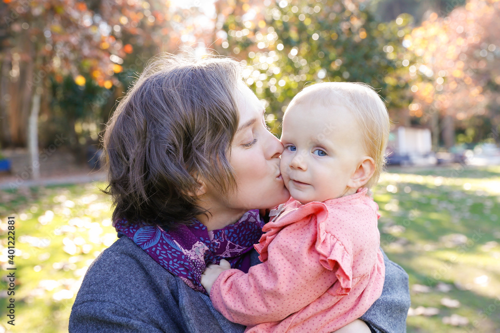 Happy mother kissing baby girl in cheek with closed eyes. Portrait of attractive Caucasian woman walking in autumn park with adorable girl sitting on mother hands. Family and maternity concept Happy mother kissing baby girl in cheek with closed eyes. Portrait of attractive Caucasian woman walking in autumn park with adorable girl sitting on mother hands. Family and maternity concept