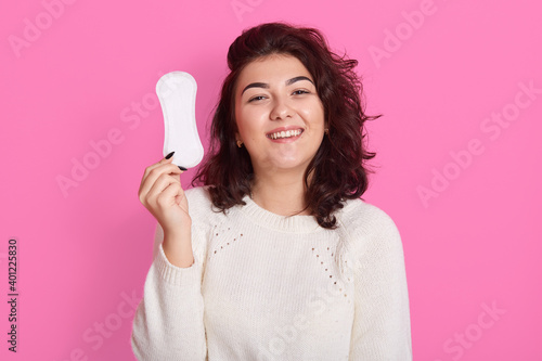 Satisfied lovely woman with dark wavy hair, holds sanitary napkin, uses hygienic product during critical days, smiles positively and looks at camera, poses isolated on pink wall.