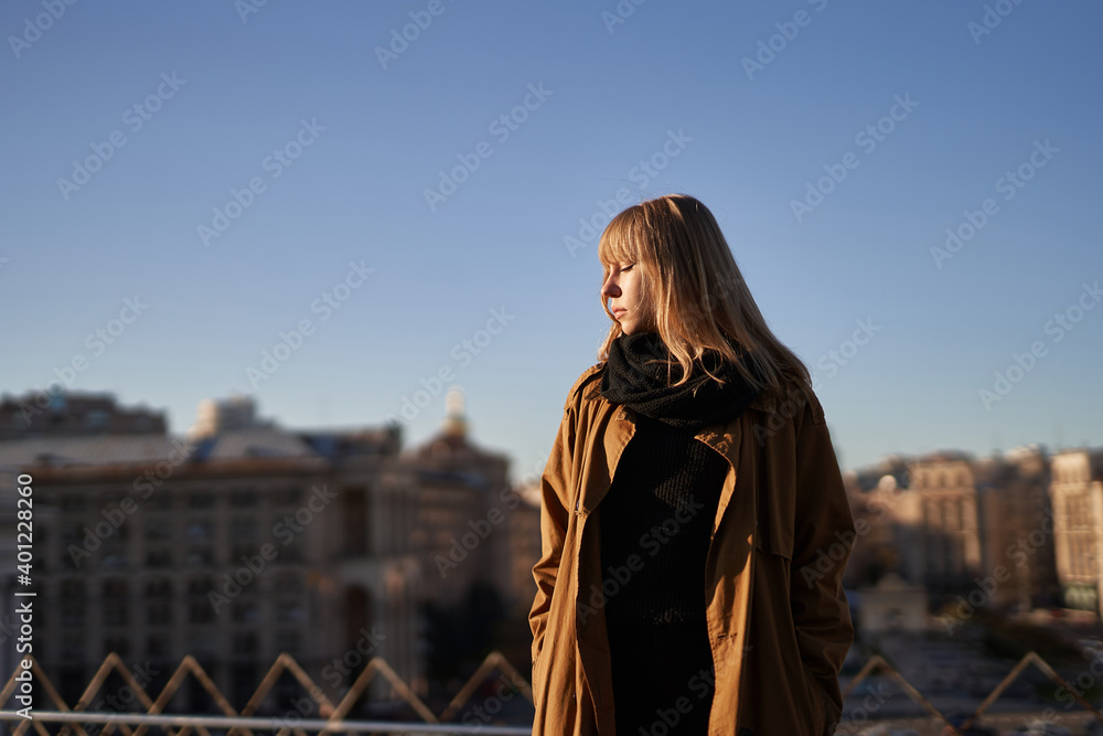 Blonde young woman in black scarf and trench standing on the european city background with blue sky. Fashion concept.
