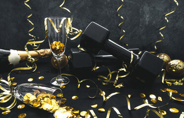 Heavy black dumbbells, champagne bottle, glasses with confetti, golden decorations for New Year's Eve celebration party. Healthy fitness lifestyle composition. Gym, workout New Year's resolution.