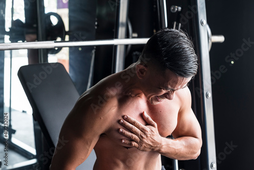 An asian male bodybuilder suffers a torn pectoral muscle while doing heavy incline bench presses on a Smith Machine at a gym or fitness center. A common workout injury