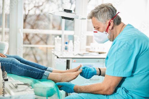 Professional doctor podiatrist in medical mask examining feet and nail while patient sitting in medical chair