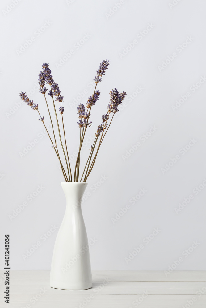 Dried lavender flowers in white vase on wooden surface over light background. Interior floral composition.