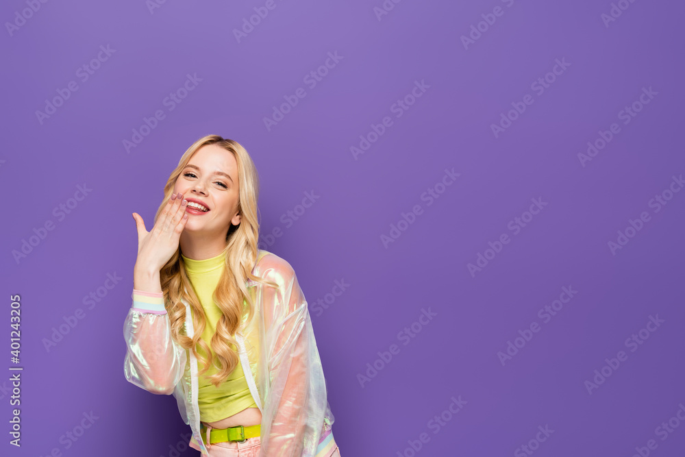 smiling blonde young woman in colorful outfit with hand near face on purple background. smiling blonde young woman in colorful outfit with hand near face on purple background.