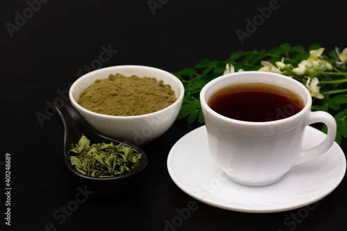 moringa tea in white cup and moringa powder in white bowl with green leaf and flower isolated on black background