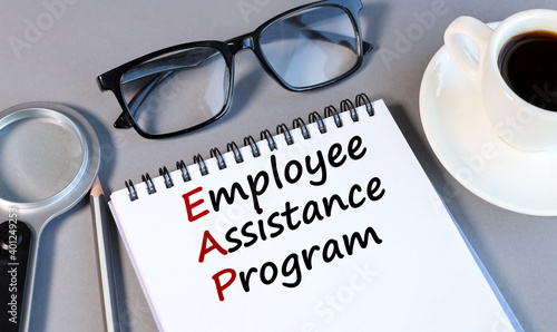 EMPLOYEE ASSISTANCE PROGRAM, text on white notepad paper on a gray background near a magnifying glass and a CUP OF COFFEE photo