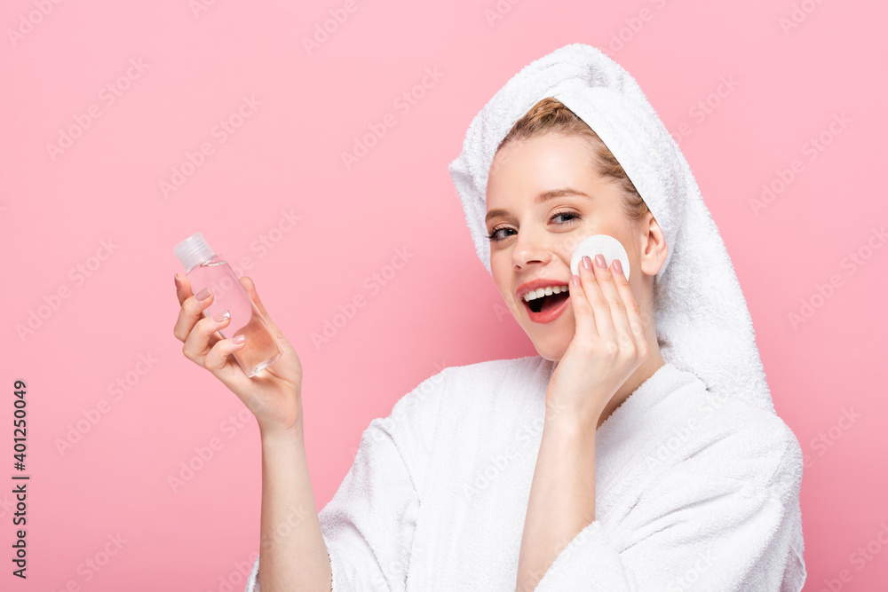 happy young woman in bathrobe with towel on head cleansing face with micellar water and cotton pad isolated on pink. happy young woman in bathrobe with towel on head cleansing face with micellar water and cotton pad isolated on pink.