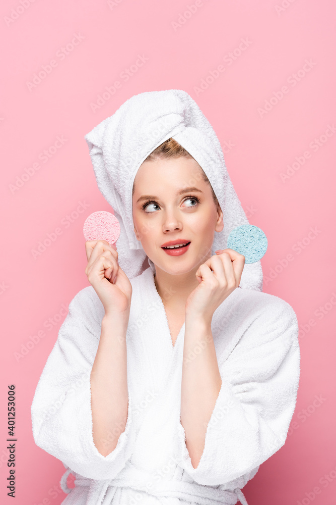 dreamy young woman in bathrobe with towel on head holding facial sponges isolated on pink. dreamy young woman in bathrobe with towel on head holding facial sponges isolated on pink.