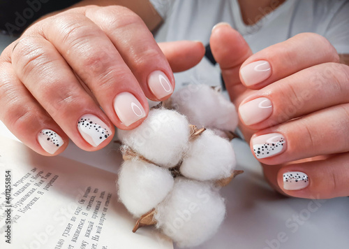 Soft pink gel polish with a design of black dots on the background of the text of the book. Women's hands with pastel colored nails hold a sprig of cotton. Milk manicure with black and white design.