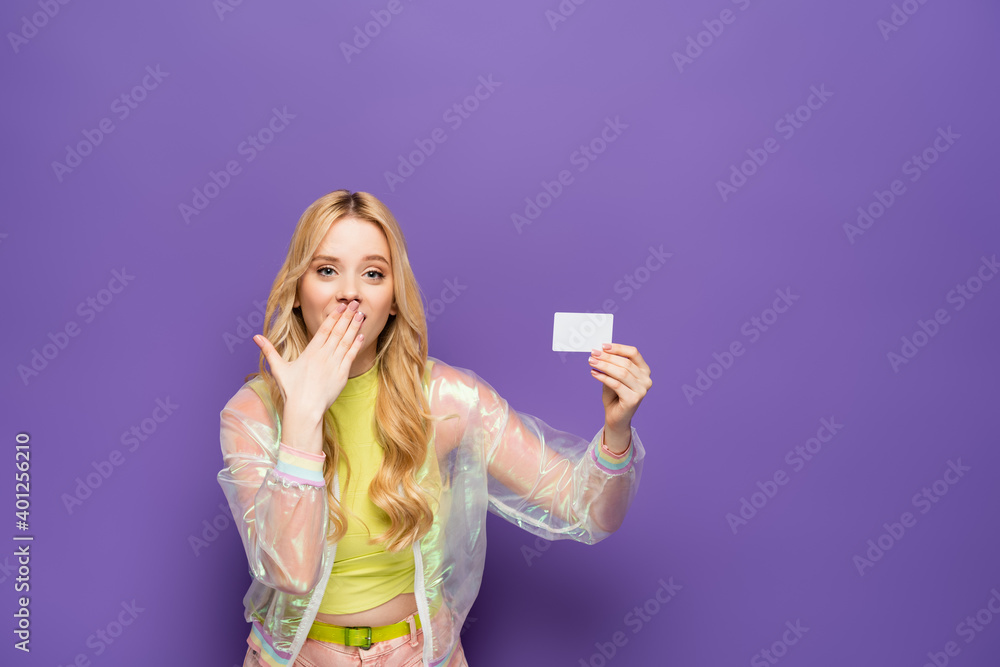 shocked blonde young woman in colorful outfit holding blank card on purple background.