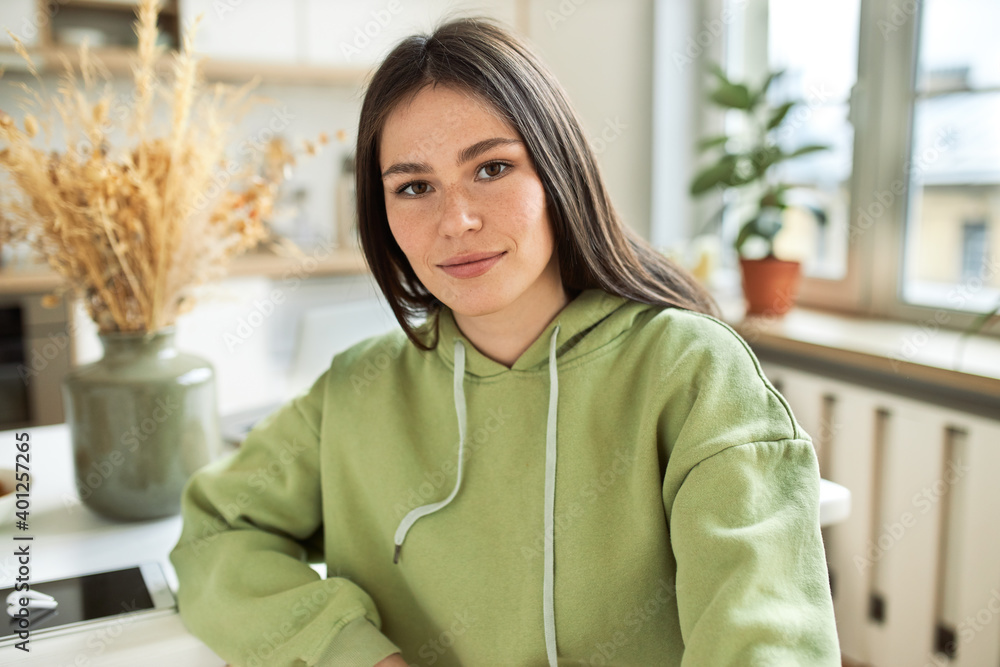 Leisure, free time and electronic devices. Indoor image of young brunette female in stylish hoodie sitting at dining table with generic cell phone and wireless earplugs, using gadgets for work Leisure, free time and electronic devices. Indoor image of young brunette female in stylish hoodie sitting at dining table with generic cell phone and wireless earplugs, using gadgets for work