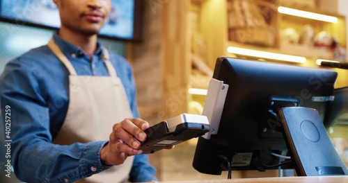 Close up of African American male seller in face mask and protective black gloves selling baked fresh bread in bakery shop. Client paying with credit card on device buying baking. Business concept