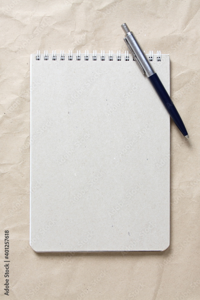 Gray notepad with white coiled spring and pen on a background of beige crumpled craft paper Gray notepad with white coiled spring and pen on a background of beige crumpled craft paper