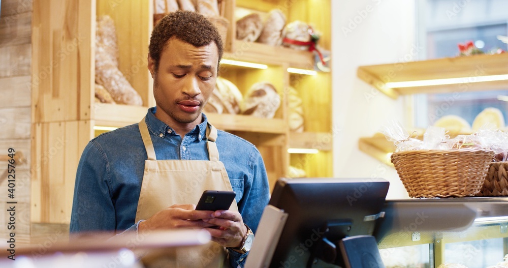 Close up of African American young handsome male worker in apron stands in bakery shop, texting and typing on smartphone. Man seller browsing online on cellphone. Business concept. Retail industry