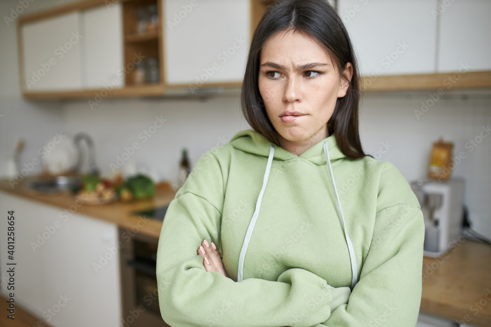 Negative human feelings and reactions. Indoor shot of frustrated stubborn teenage girl in hoodie biting lips, posing i kitchen with arms crossed, expressing disagreement, does not want to do housework Negative human feelings and reactions. Indoor shot of frustrated stubborn teenage girl in hoodie biting lips, posing i kitchen with arms crossed, expressing disagreement, does not want to do housework