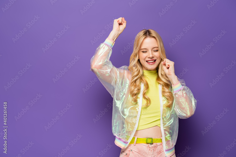 happy blonde young woman in colorful outfit showing yeah gesture on purple background. happy blonde young woman in colorful outfit showing yeah gesture on purple background.